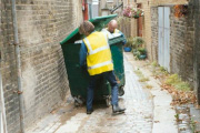 Two people in hi-viz moving a large overfull wheelie bin along a rough cobbled allyway